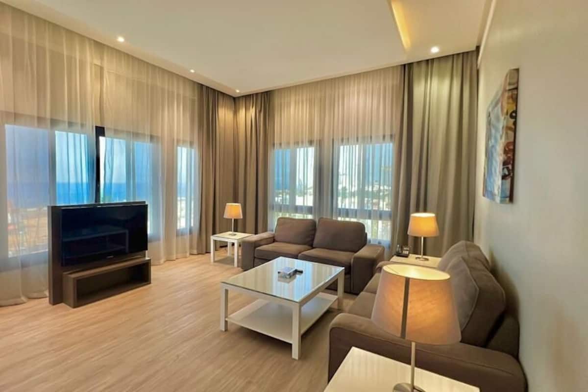 A modern hotel room with a flat-screen TV, two sofas, a glass coffee table, and a large window with curtains.