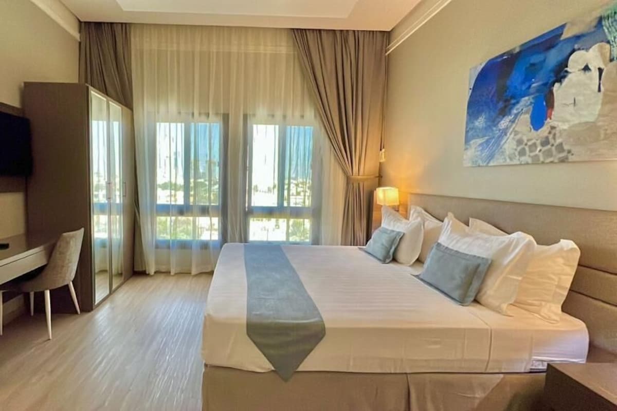 A hotel room with a bed, desk, chair, TV, and a large window with curtains.