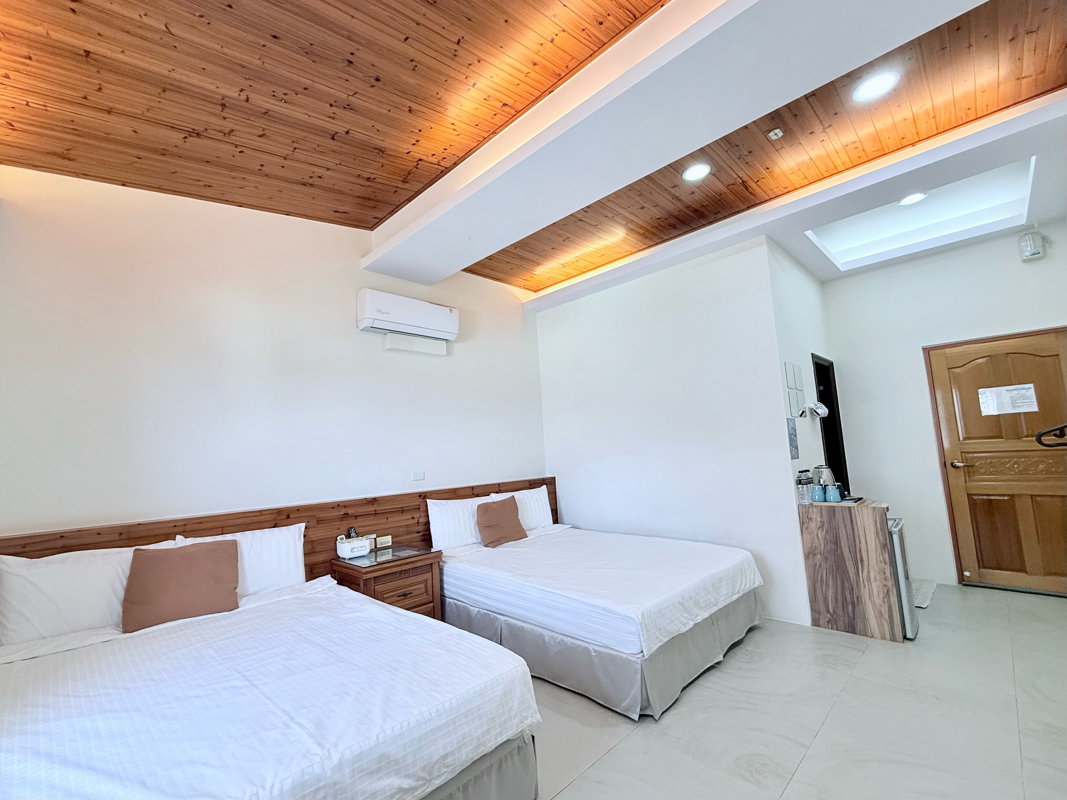 A hotel room with two beds, a wooden ceiling, an air conditioning unit, a wooden door, and a small table with toiletries.