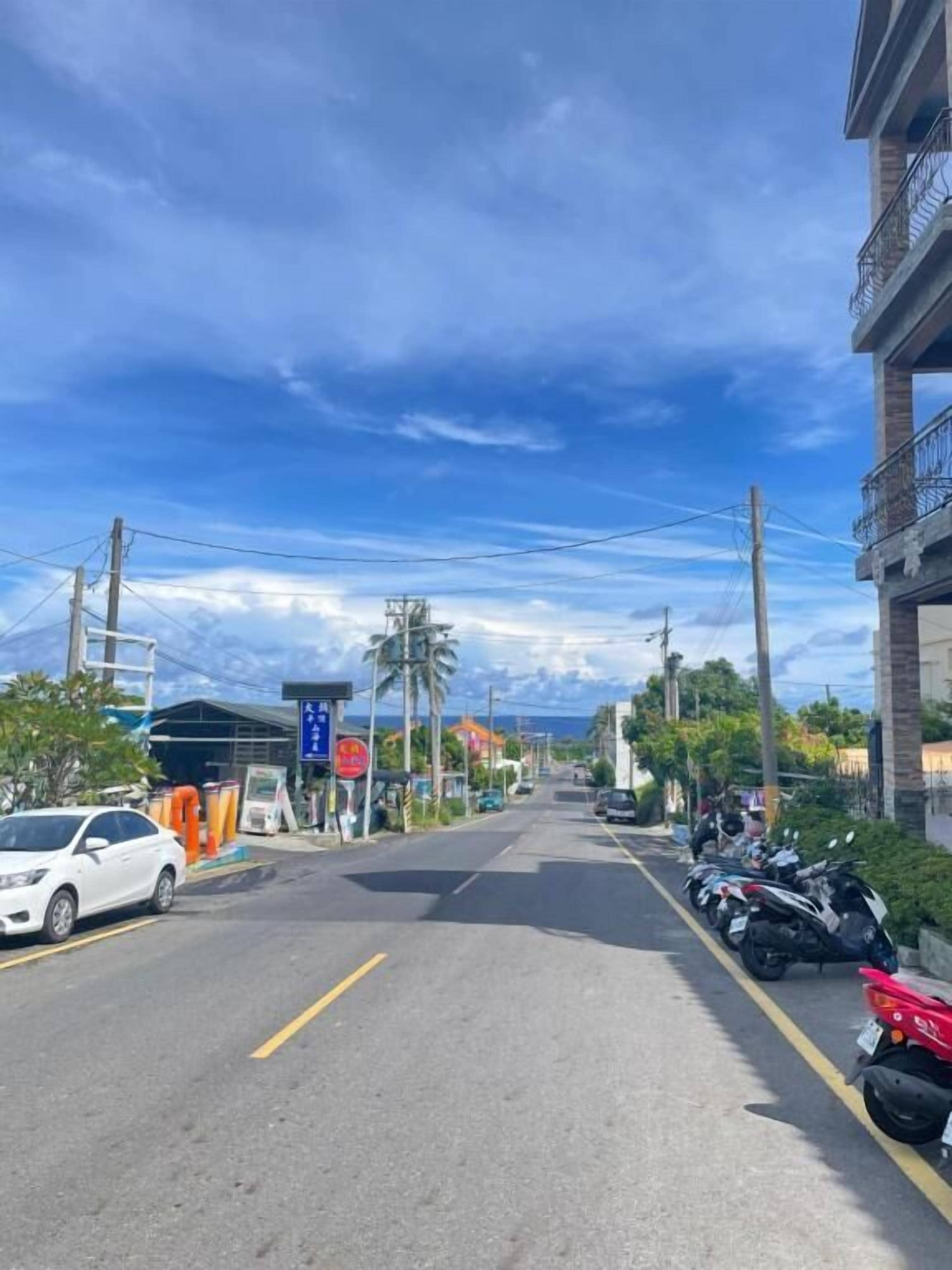 Eine Straße mit abgestellten Motorrädern und einem weißen Auto, mit Gebäuden an beiden Seiten und Stromleitungen darüber.