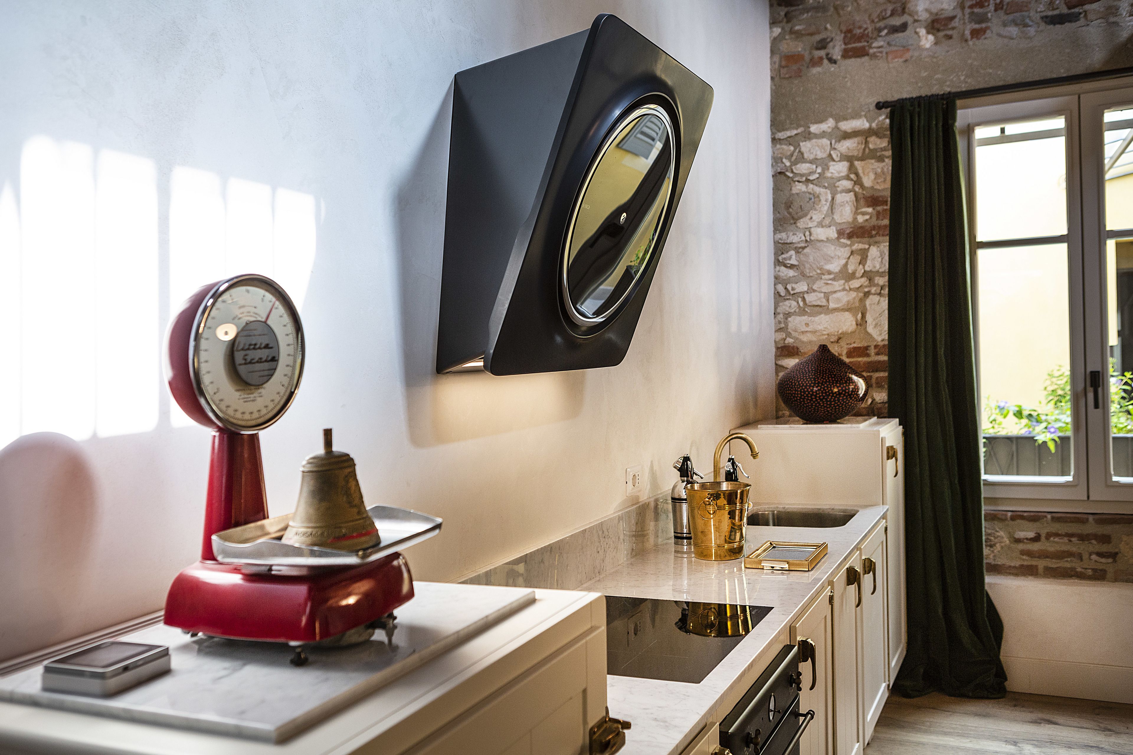 Una cocina con pared de piedra, una balanza roja, un reloj negro y una ventana con cortinas.