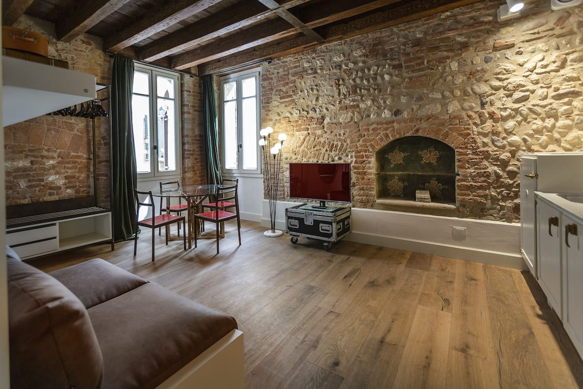 A rustic room with a stone wall, wooden floor, a fireplace, a sofa, a dining table with chairs, and a small cabinet.