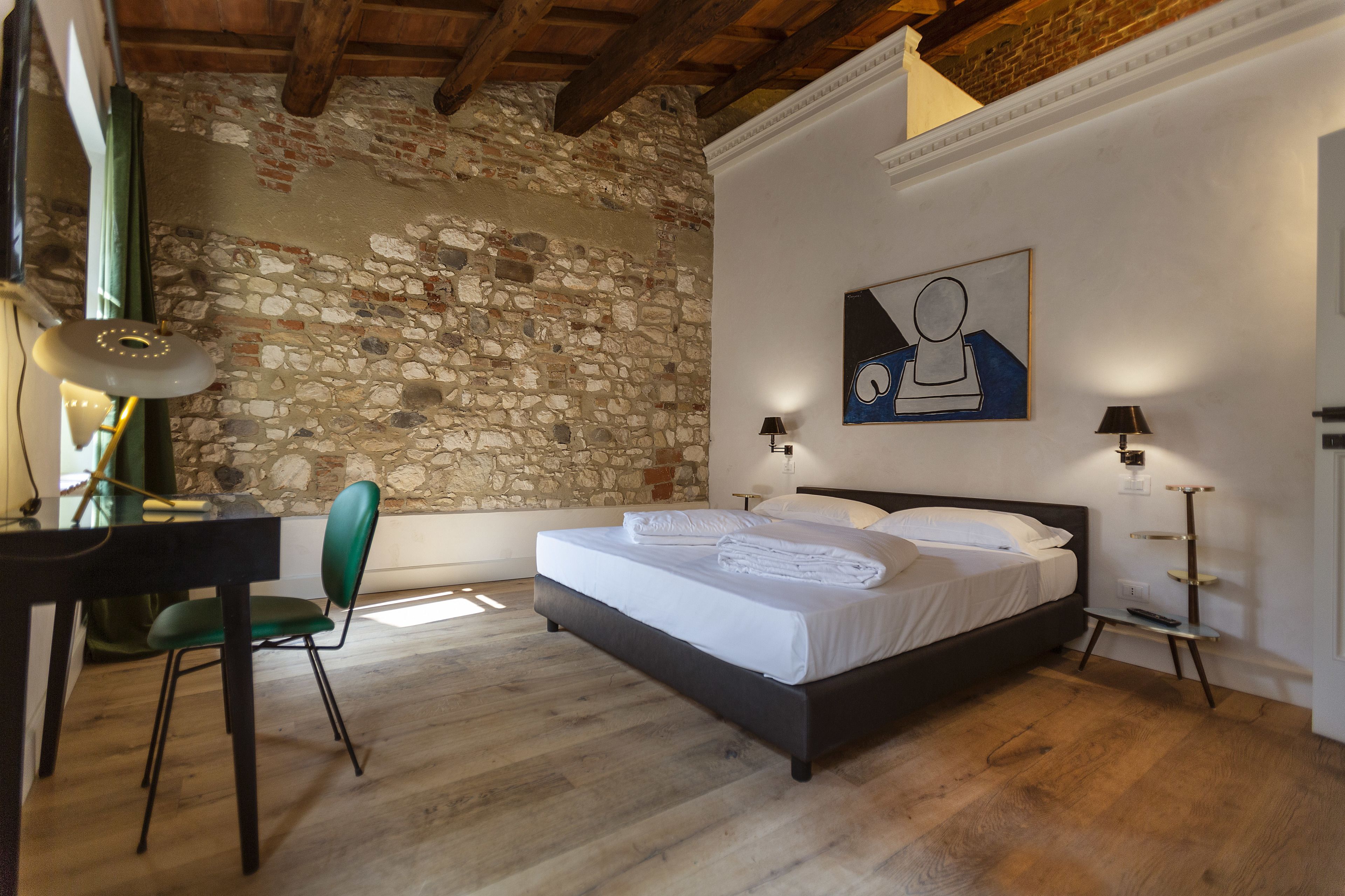 A bedroom with a stone wall, a bed with white linens, a desk with a chair, and two bedside lamps.