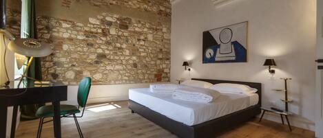 A bedroom with a stone wall, a bed with white linens, a desk with a chair, and two bedside lamps.