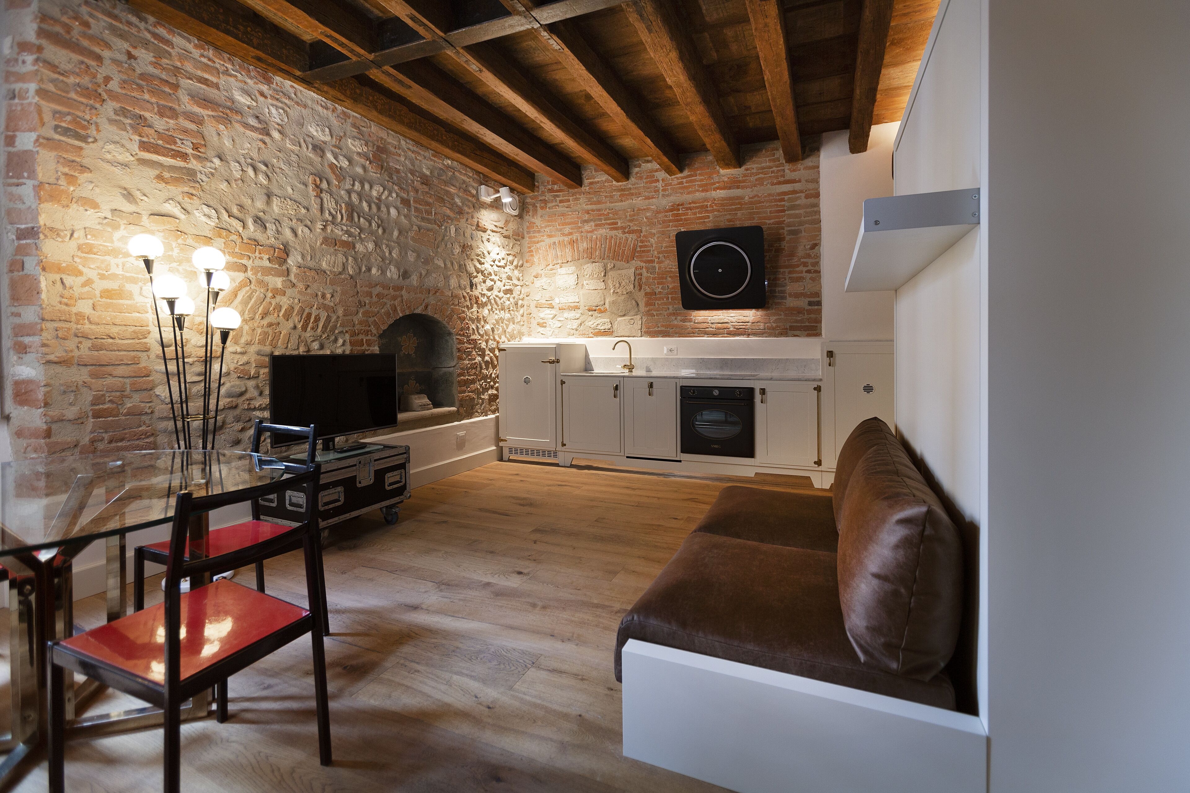 A rustic living room with exposed brick walls, a wooden ceiling, a glass-top dining table with chairs, a brown sofa, and a kitchen area with white cabinets and a black speaker.