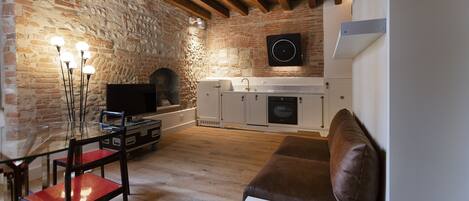 A rustic living room with exposed brick walls, a wooden ceiling, a glass-top dining table with chairs, a brown sofa, and a kitchen area with white cabinets and a black speaker.