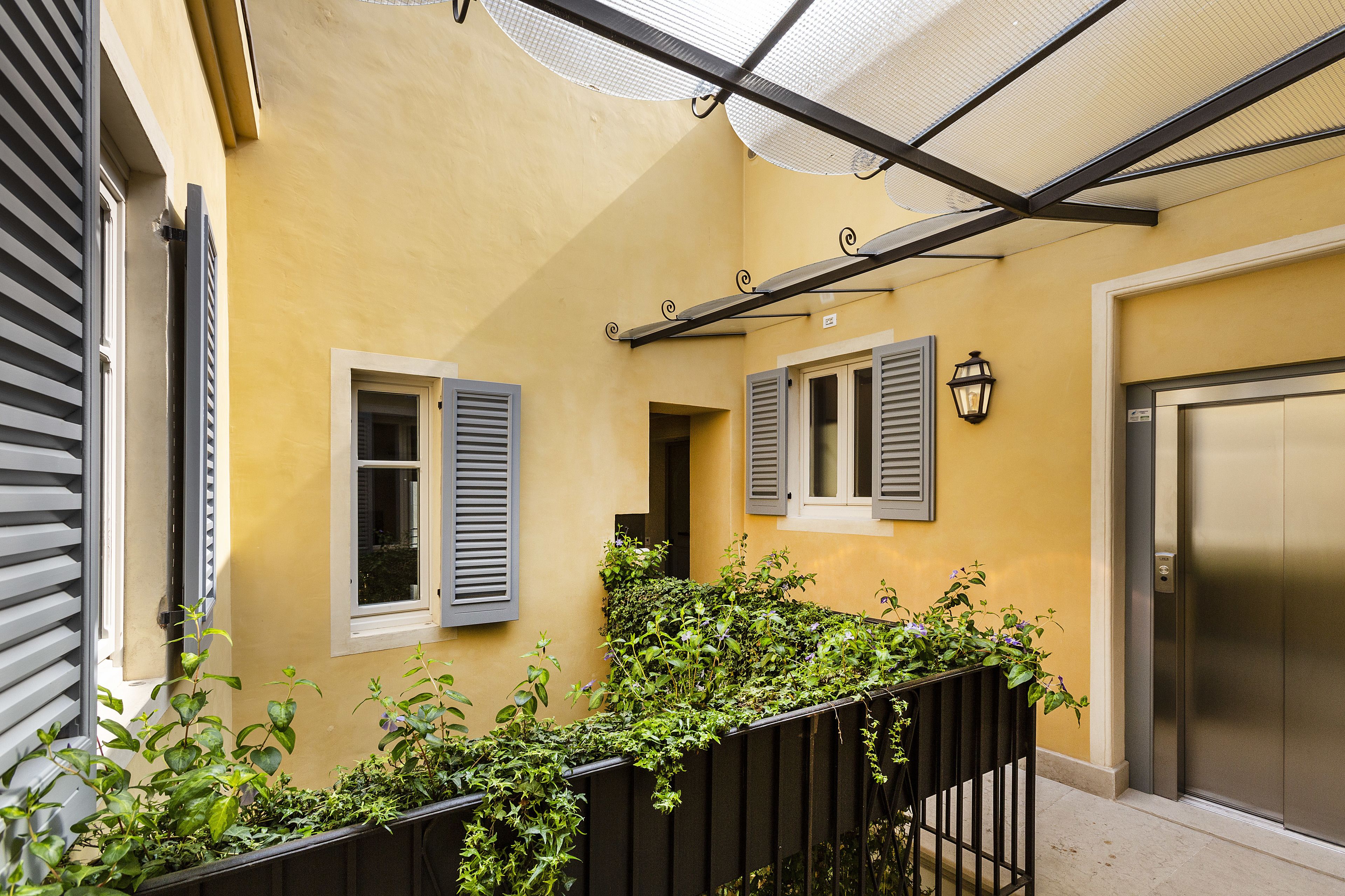 A yellow building with a metal elevator, a window with shutters, and a metal railing with greenery.