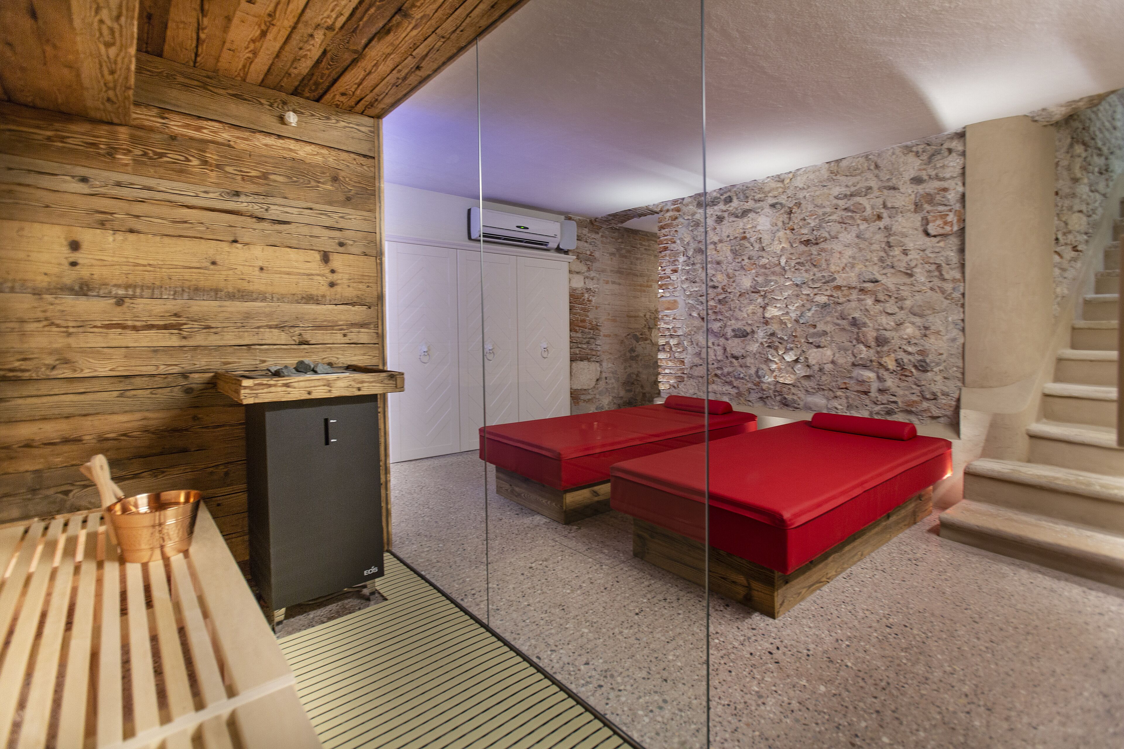 A sauna room with wooden walls, a stone wall, a wooden bench, and two red massage beds.