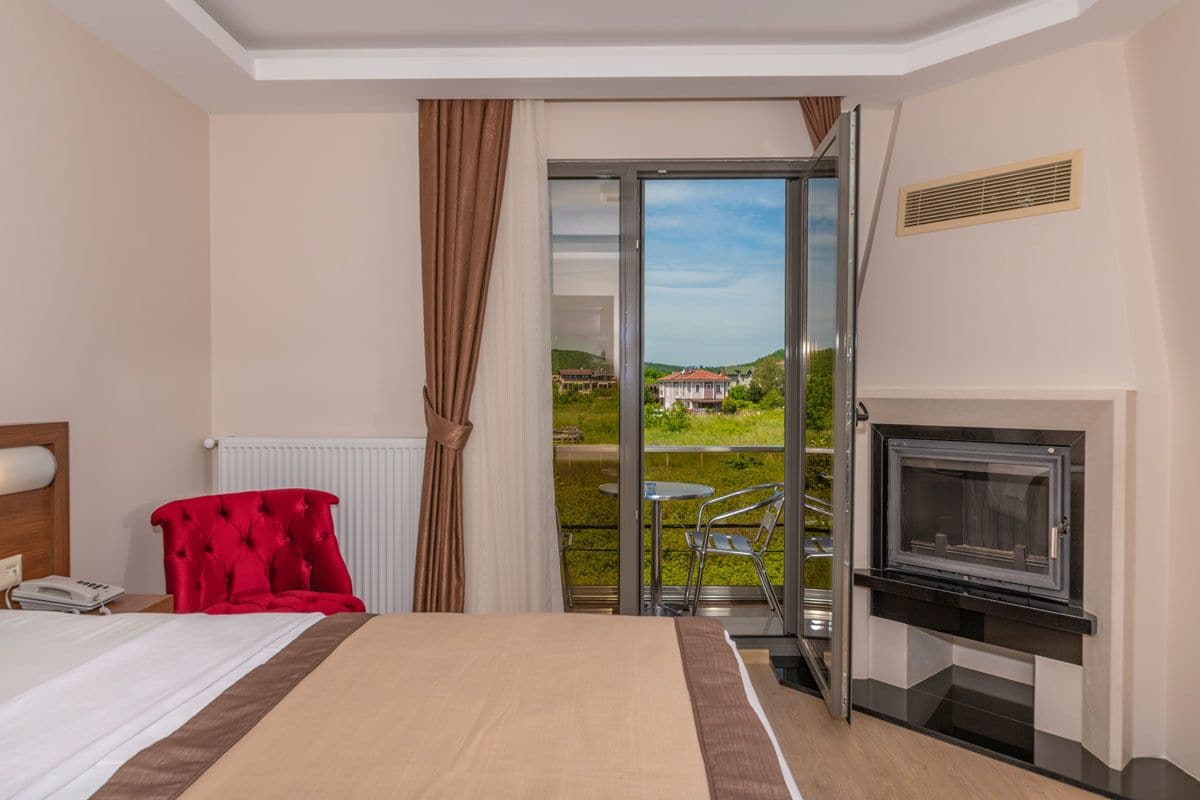 Habitación de hotel con cama, un sillón rojo, televisor y puerta corrediza de vidrio que da a un balcón con vista a una casa y follaje.