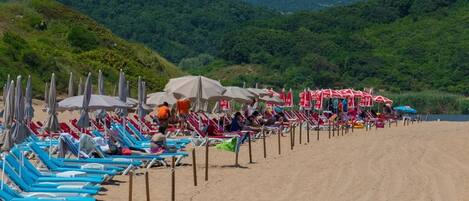 A beach with rows of sun loungers and umbrellas, a sandy beach, and a forested hillside in the background.