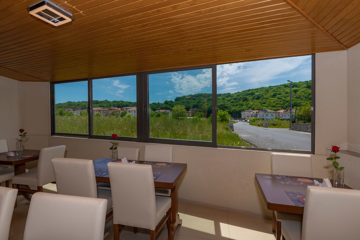A dining area with wooden tables and chairs, large windows with a view of a road and greenery.