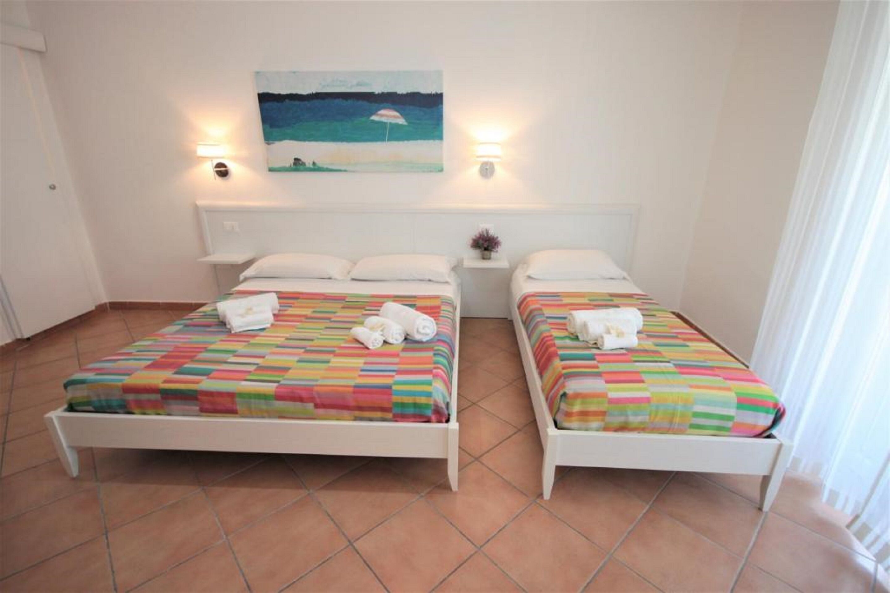Une chambre double avec deux lits, chacun recouvert d’une courtepointe rayée aux couleurs vives et agrémenté de coussins blancs. Un tableau est accroché au mur au-dessus des lits, et deux lampes sont fixées au mur.