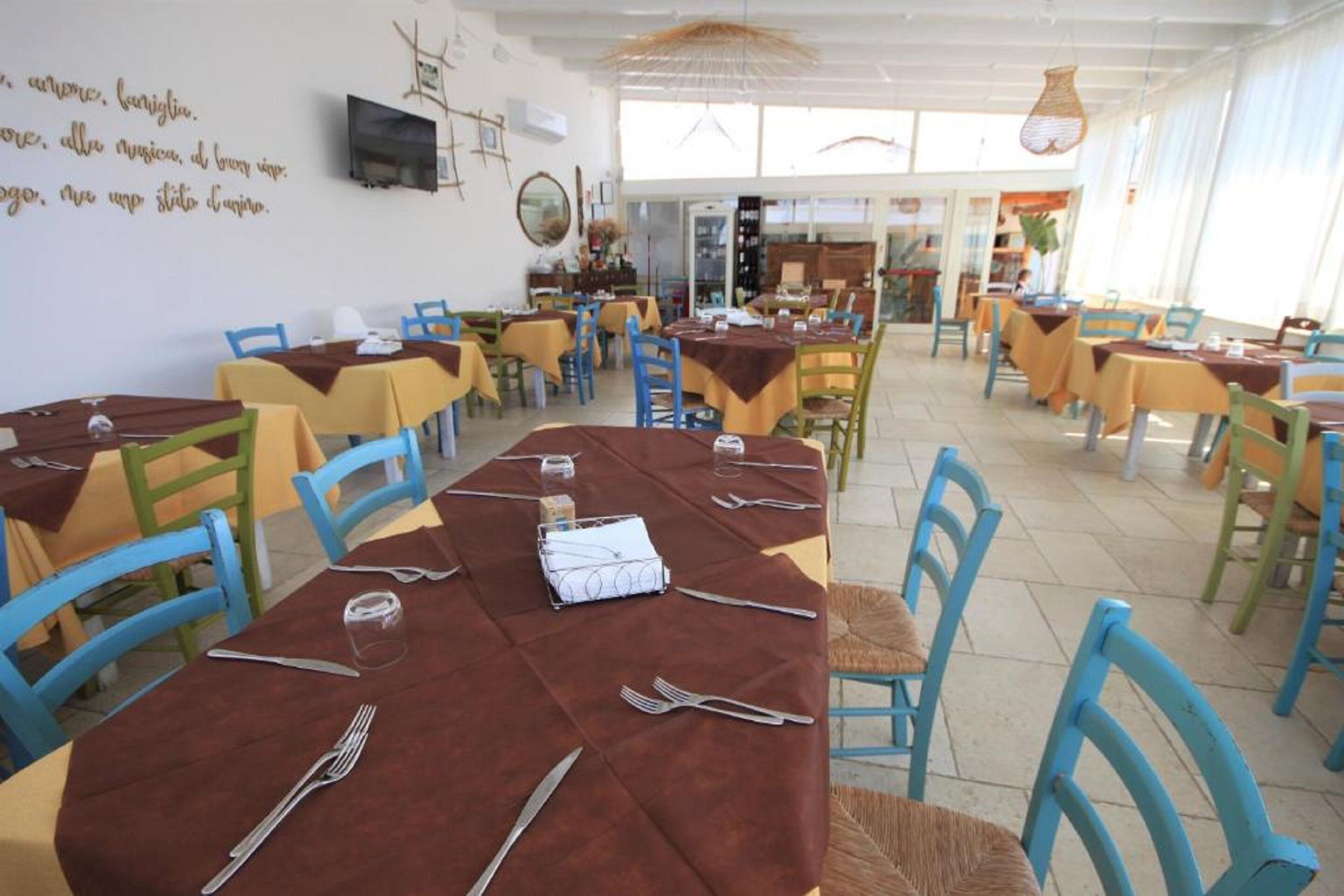 Una sala ristorante con diversi tavoli e sedie, alcuni con tovaglie marroni e altri gialle. Sui tavoli si notano bicchieri, forchette e coltelli. Le pareti sono decorate e c'è una televisione appesa.