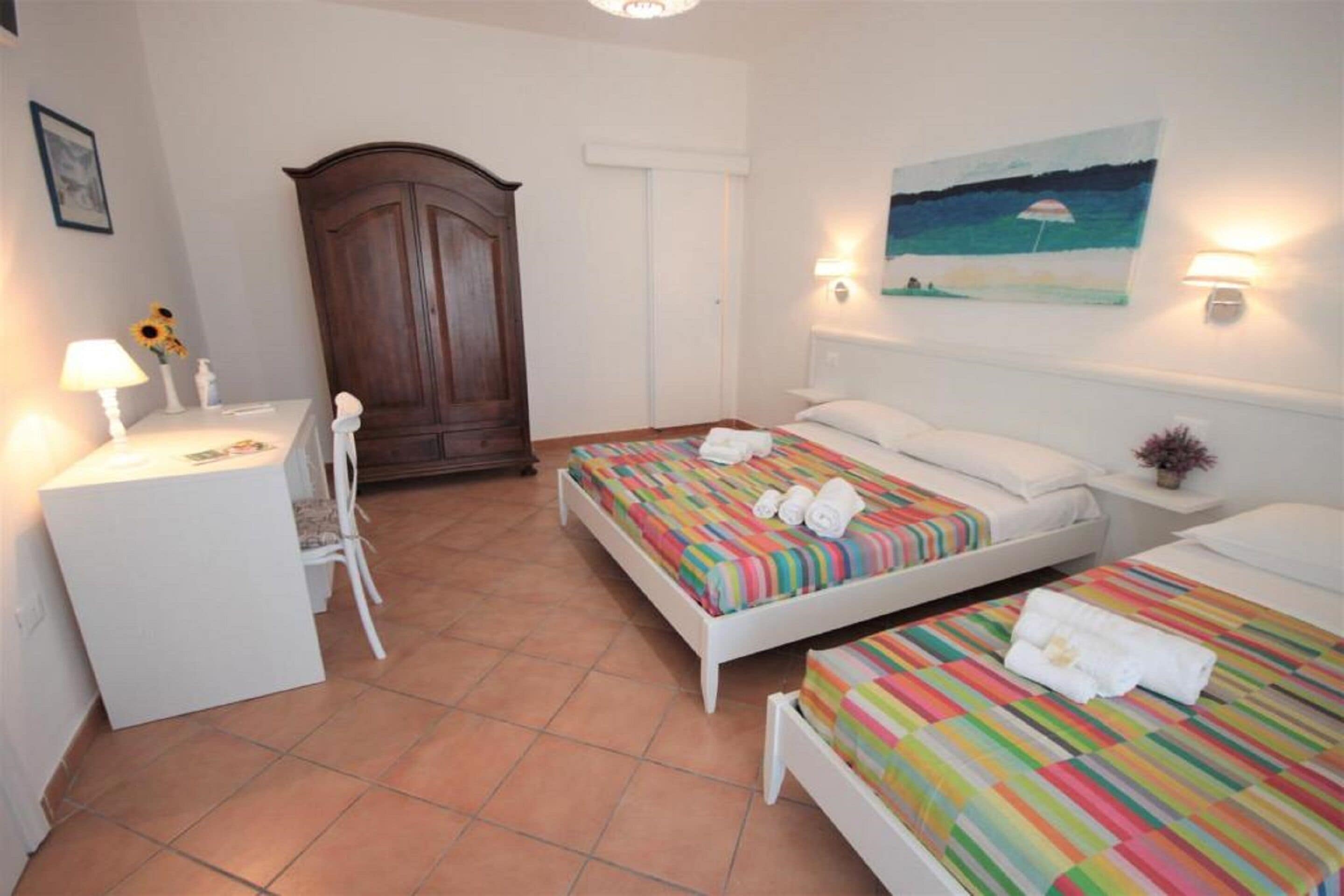 A hotel room with two beds, a desk, and a chair. There is a wardrobe and a painting on the wall.