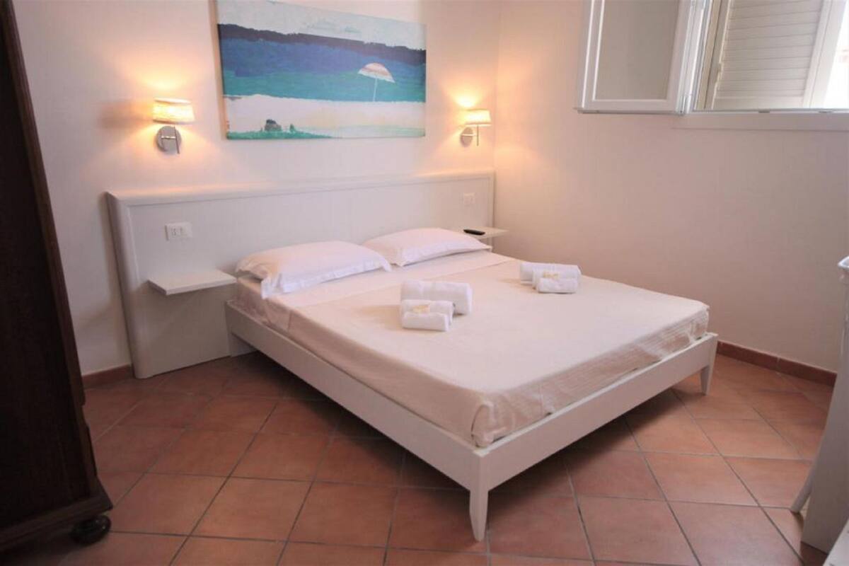 A bedroom with a bed, two bedside tables, a painting on the wall, and a window with blinds.