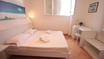 A clean, well-lit bedroom with a bed, bedside tables, a small desk, and a window with shutters.
