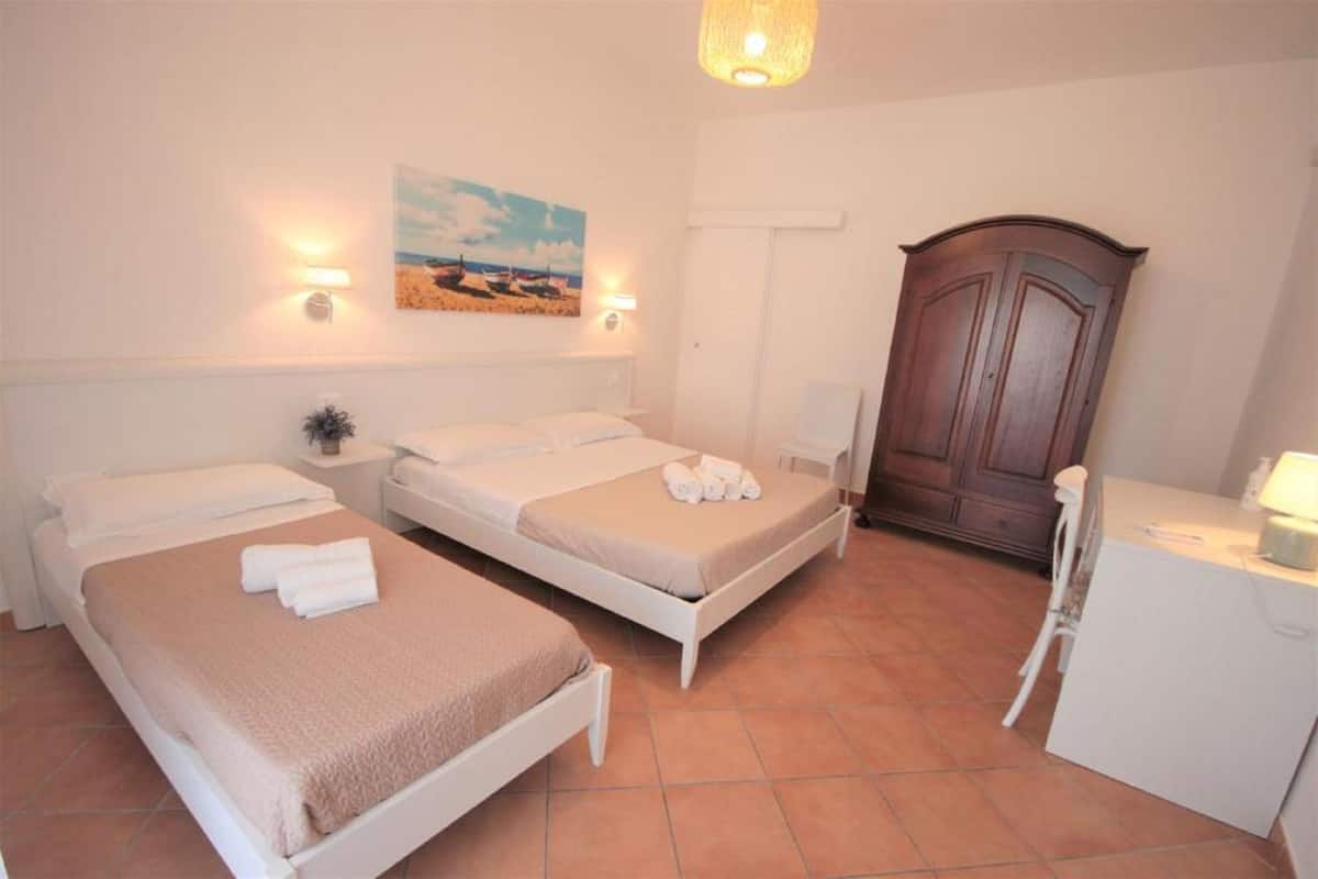 A room with two beds, a wooden wardrobe, a desk with a chair, and a painting on the wall.