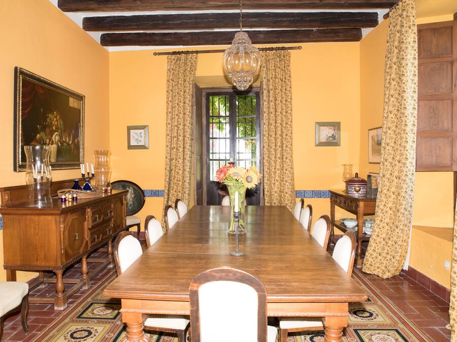 A dining room with a long wooden table, chairs, a chandelier, and a window with curtains.