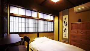 Una tradizionale stanza giapponese con pavimento in tatami, una bassa scrivania in legno, un letto con biancheria da letto bianca e una finestra con paravento scorrevole shoji.