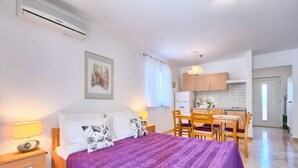 A bedroom with a bed, bedside table, and a nightstand. There is a window with curtains, a wall-mounted air conditioner, and a framed picture above the bed.