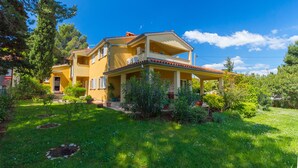 A two-story yellow house with a balcony, surrounded by greenery and a clear blue sky.