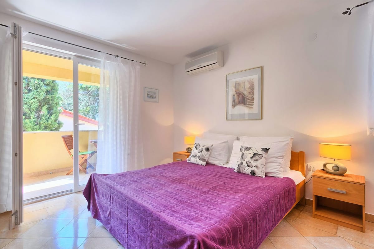 Quarto com cama, mesas de cabeceira, abajur, quadro na parede e porta de vidro deslizante para área externa.