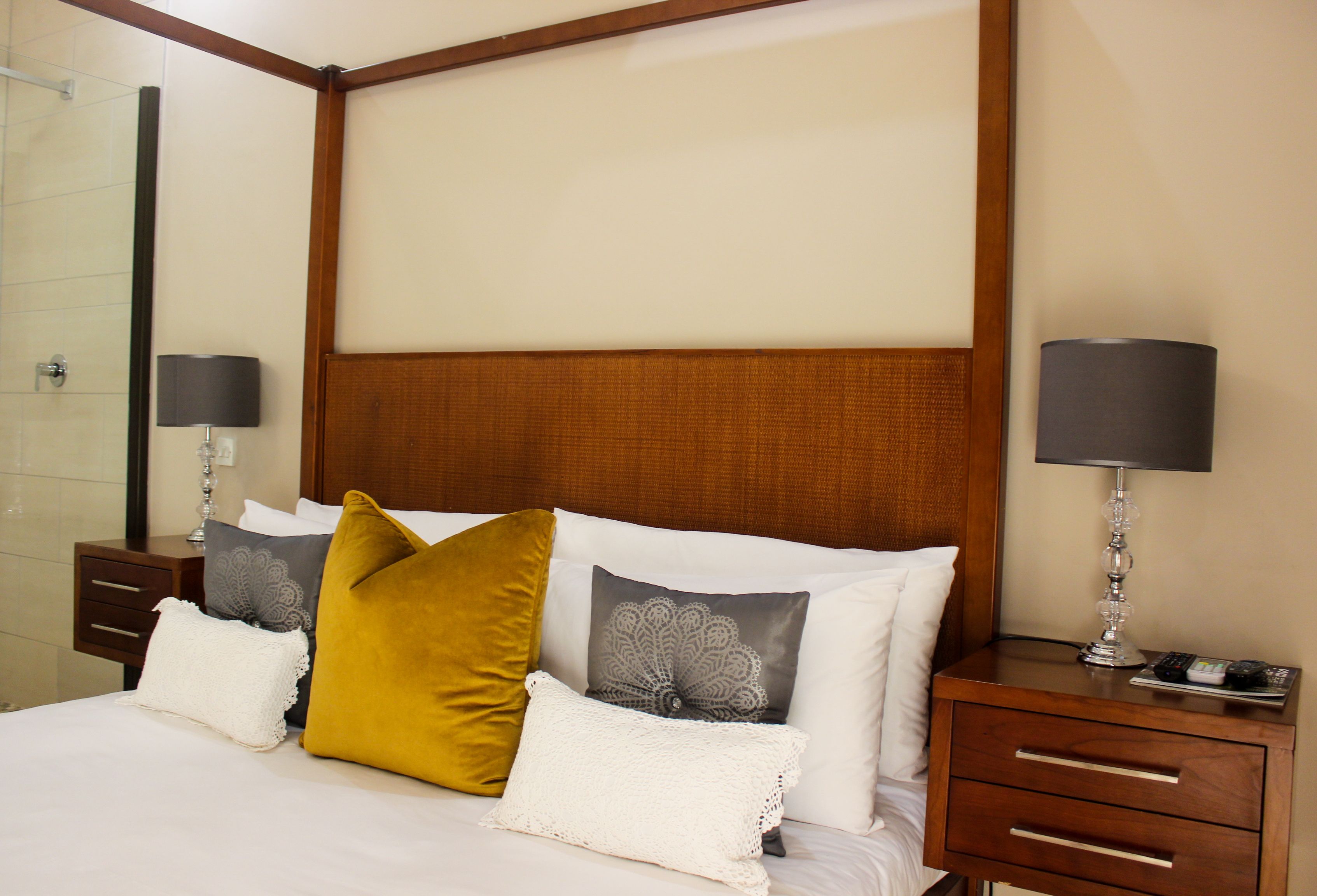 A neatly made bed with white and mustard pillows, a wooden headboard, and a bedside table with a lamp and remote control.