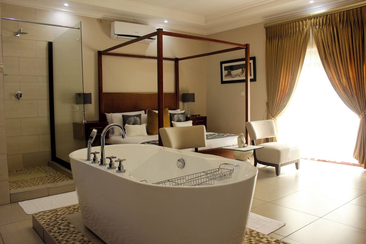 A spacious bedroom with a large bathtub, a separate shower area, a four-poster bed, and a seating area with a chair and table.
