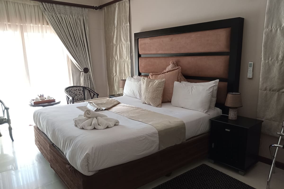 A neatly made bed with a brown headboard, a bedside table with a lamp, and a chair with a tray of breakfast items.