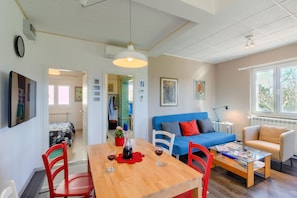 Un salon bien éclairé, avec une table à manger en bois, des chaises rouges, un canapé bleu et un fauteuil beige. Une fenêtre laisse entrer la lumière naturelle, et une porte donne accès à une autre pièce.
