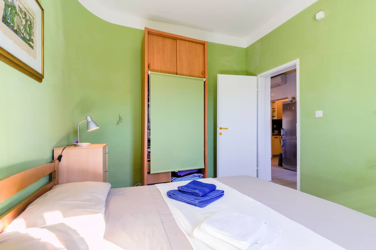 A bedroom with a bed, a nightstand, a lamp, a wardrobe, and a door leading to a kitchen.