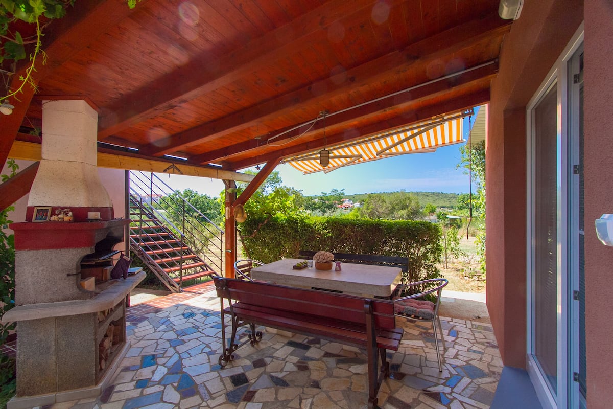 A covered patio with a stone-tiled floor, a wooden table, and a grill.