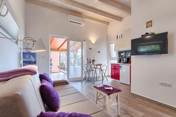 Un salon moderne avec un canapé, une petite table et un téléviseur. On y trouve également un coin cuisine avec un meuble rouge et un téléviseur fixé au mur.
