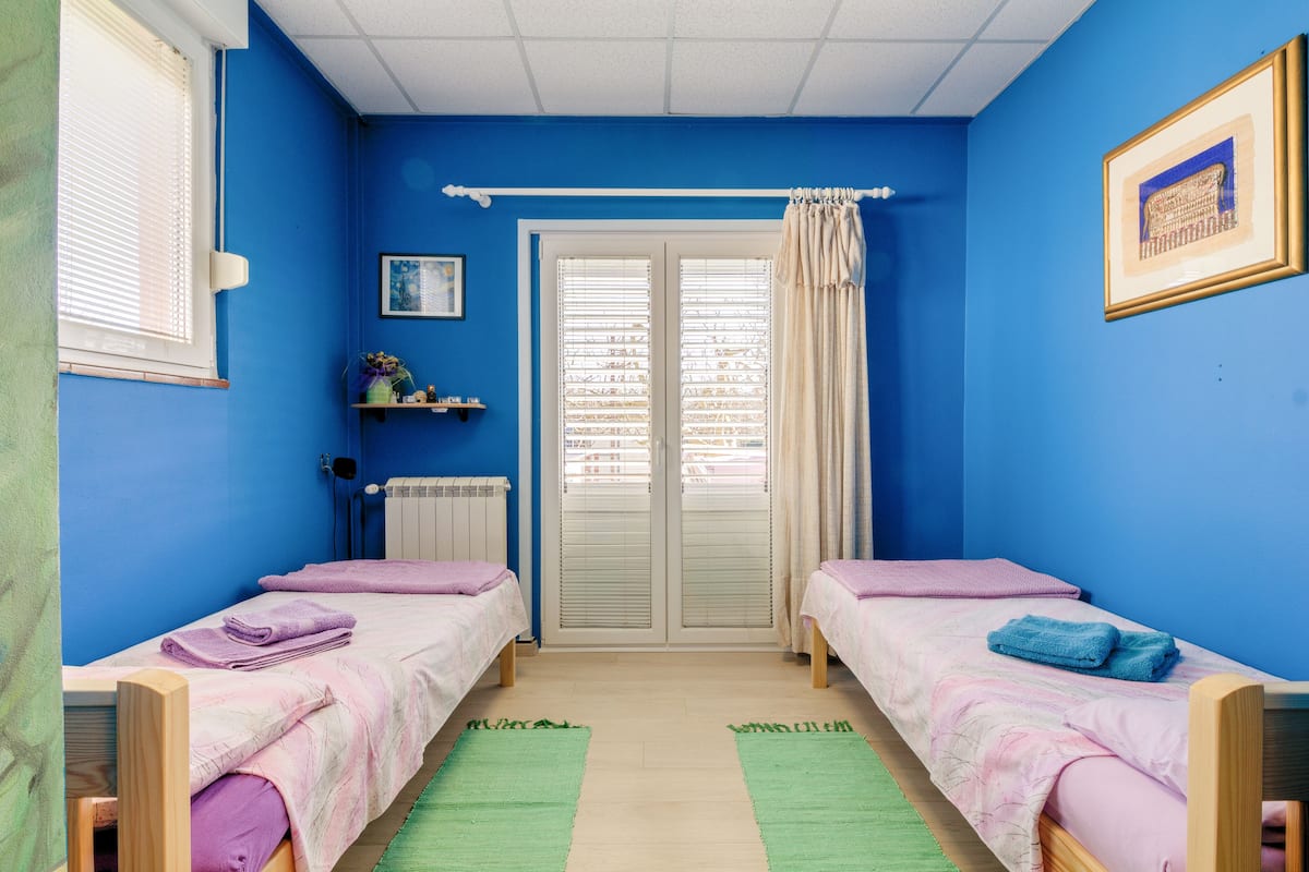A room with two beds, a radiator, a window with blinds, a door with curtains, and a framed picture on the wall.
