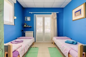 A room with two beds, a radiator, a window with blinds, a door with curtains, and a framed picture on the wall.