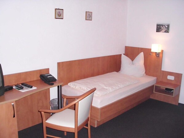 A hotel room with a bed, a desk with a telephone, a chair, and a small bedside table with a lamp. - Pension Dreiländereck (Birx)