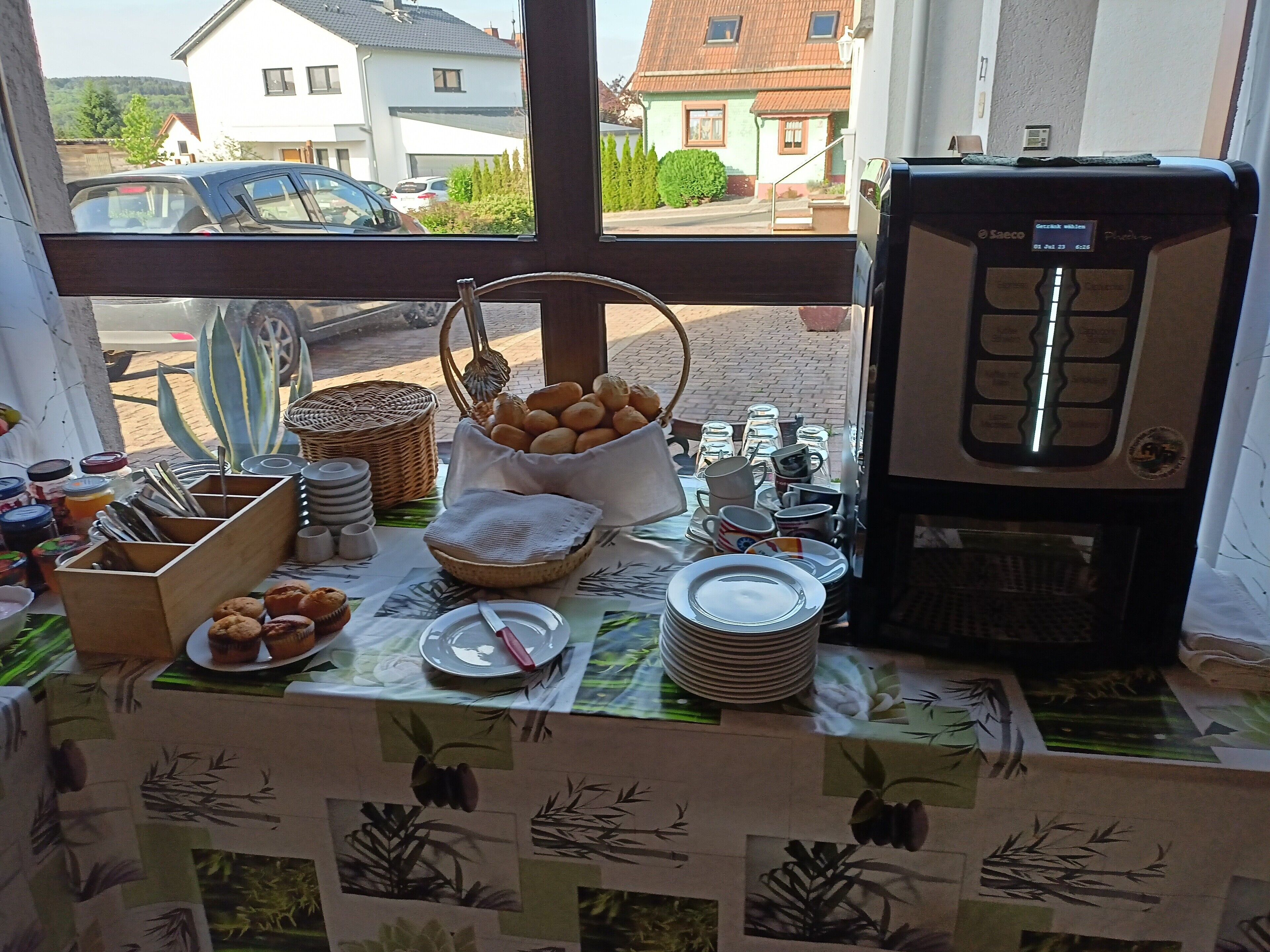 Een ontbijtbuffet met brood, gebak en bestek op een tafel, met uitzicht op huizen en geparkeerde auto's.