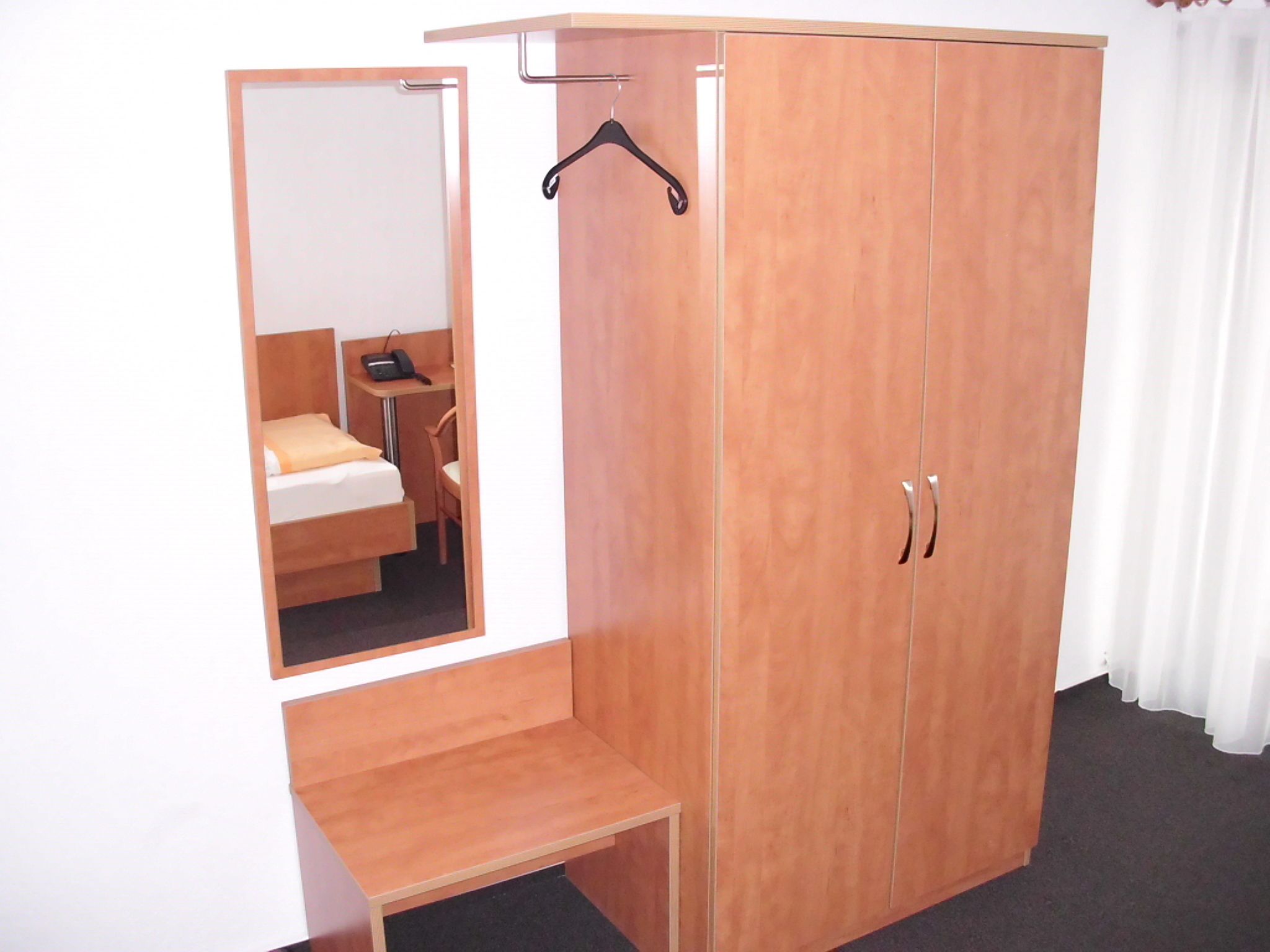 Un dormitorio con un armario de madera, una mesita de noche, un abrigo negro colgado y un espejo que refleja la habitación.