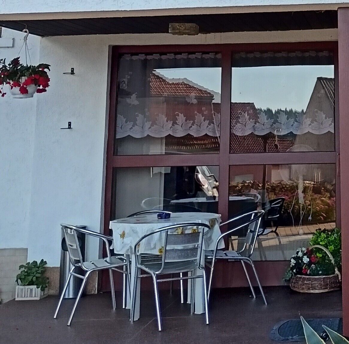 Un área de estar al aire libre con una mesa y sillas, una planta colgante y una ventana con cortinas de encaje.