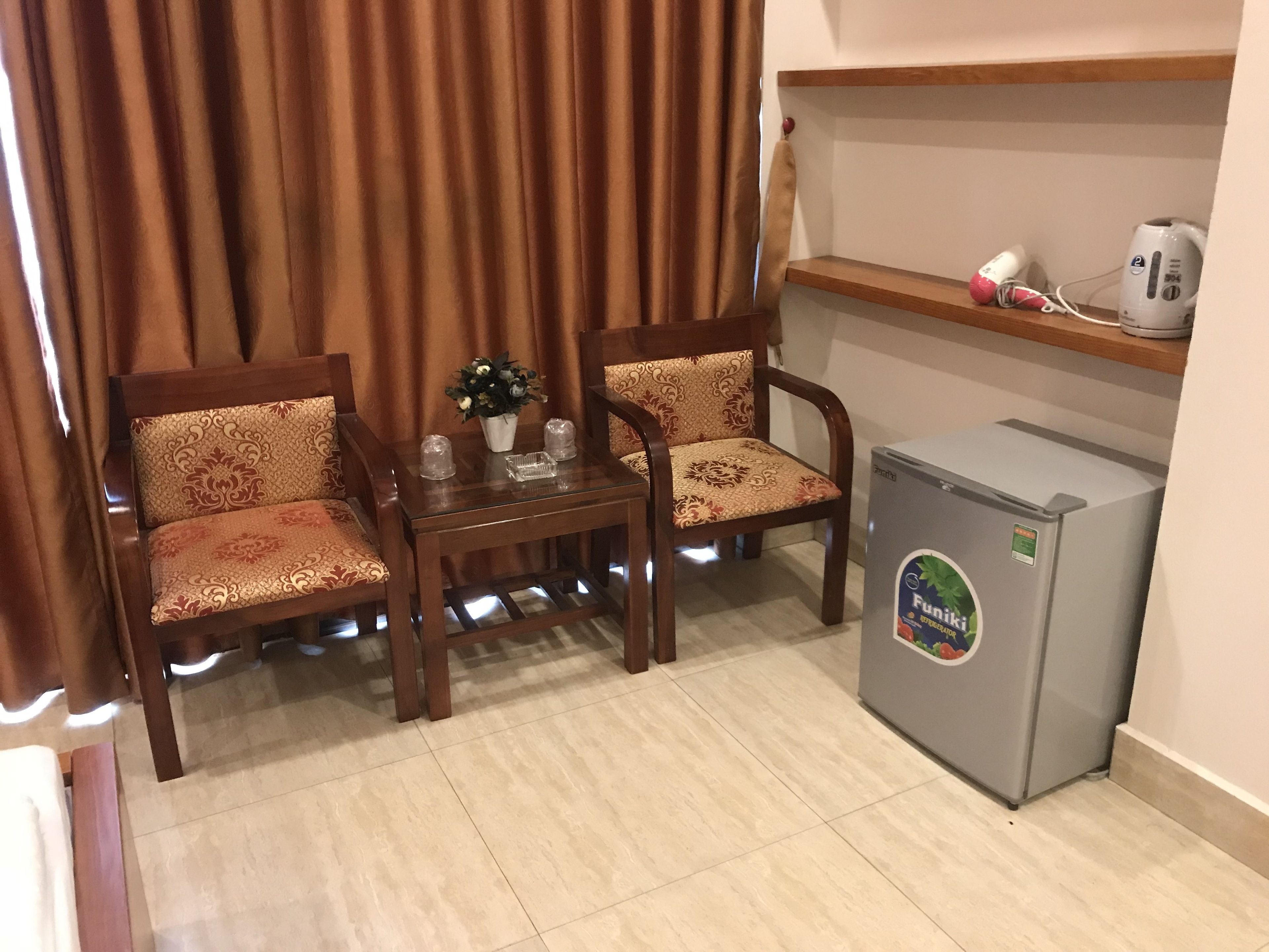 A small room with two wooden chairs with patterned cushions, a glass table with a vase and two candles, and a grey refrigerator with a sticker.
