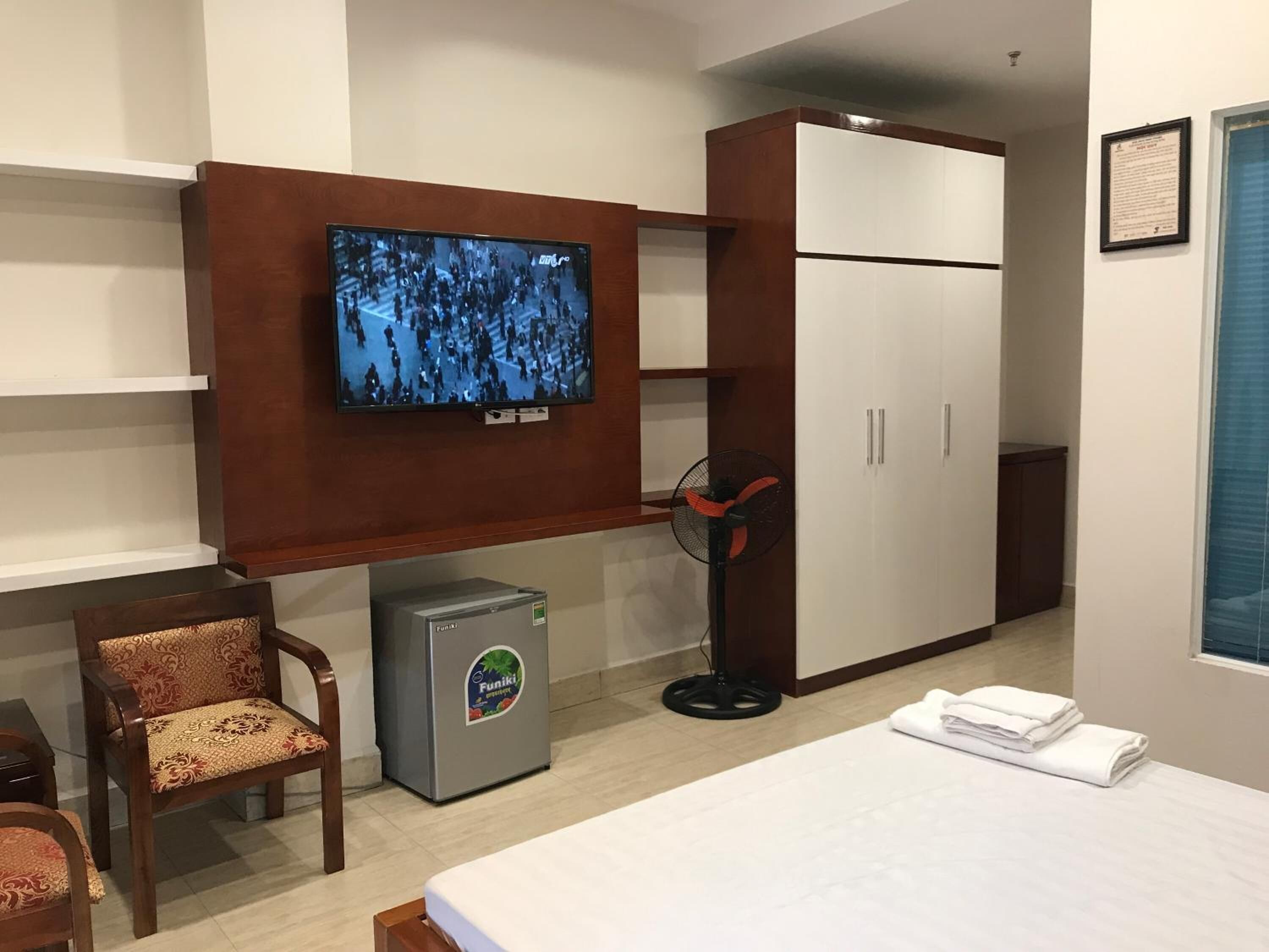 Habitación de hotel con cama, televisor montado en la pared, refrigerador pequeño, escritorio con silla y armario.