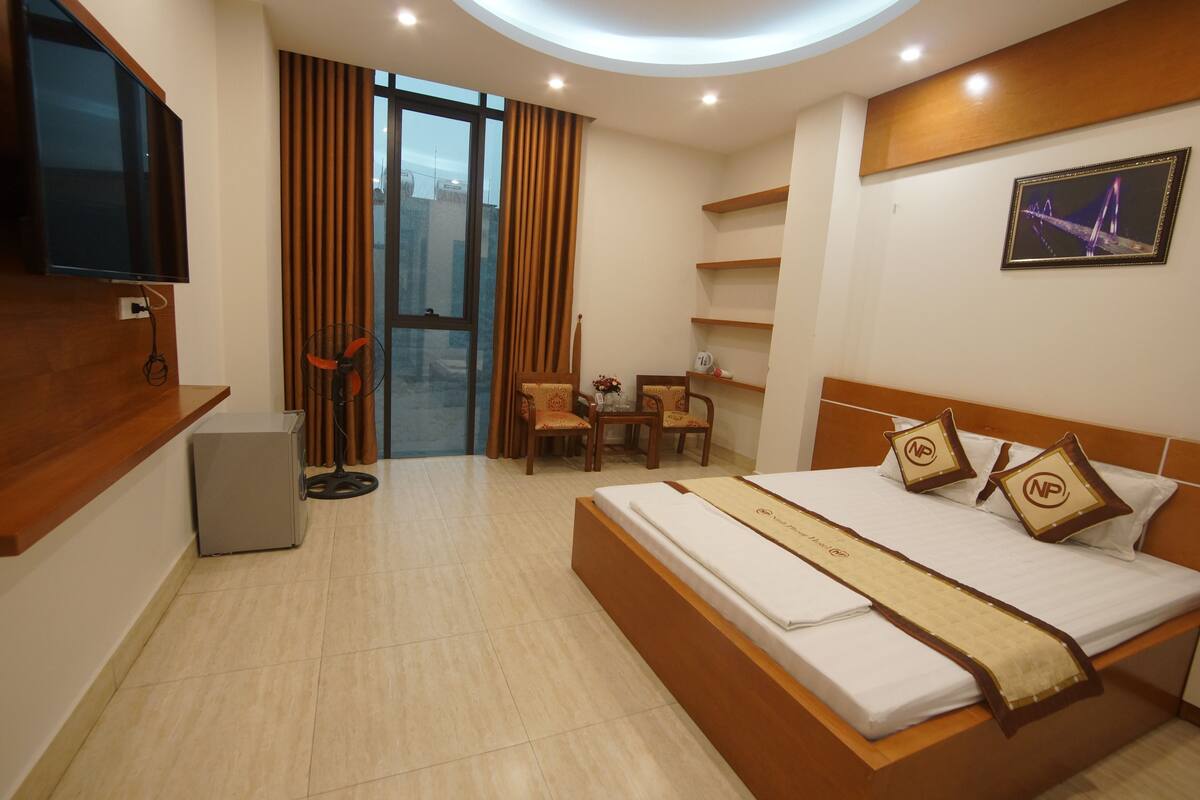 A hotel room with a bed, a desk, a chair, a TV, a small fridge, and a window with curtains.