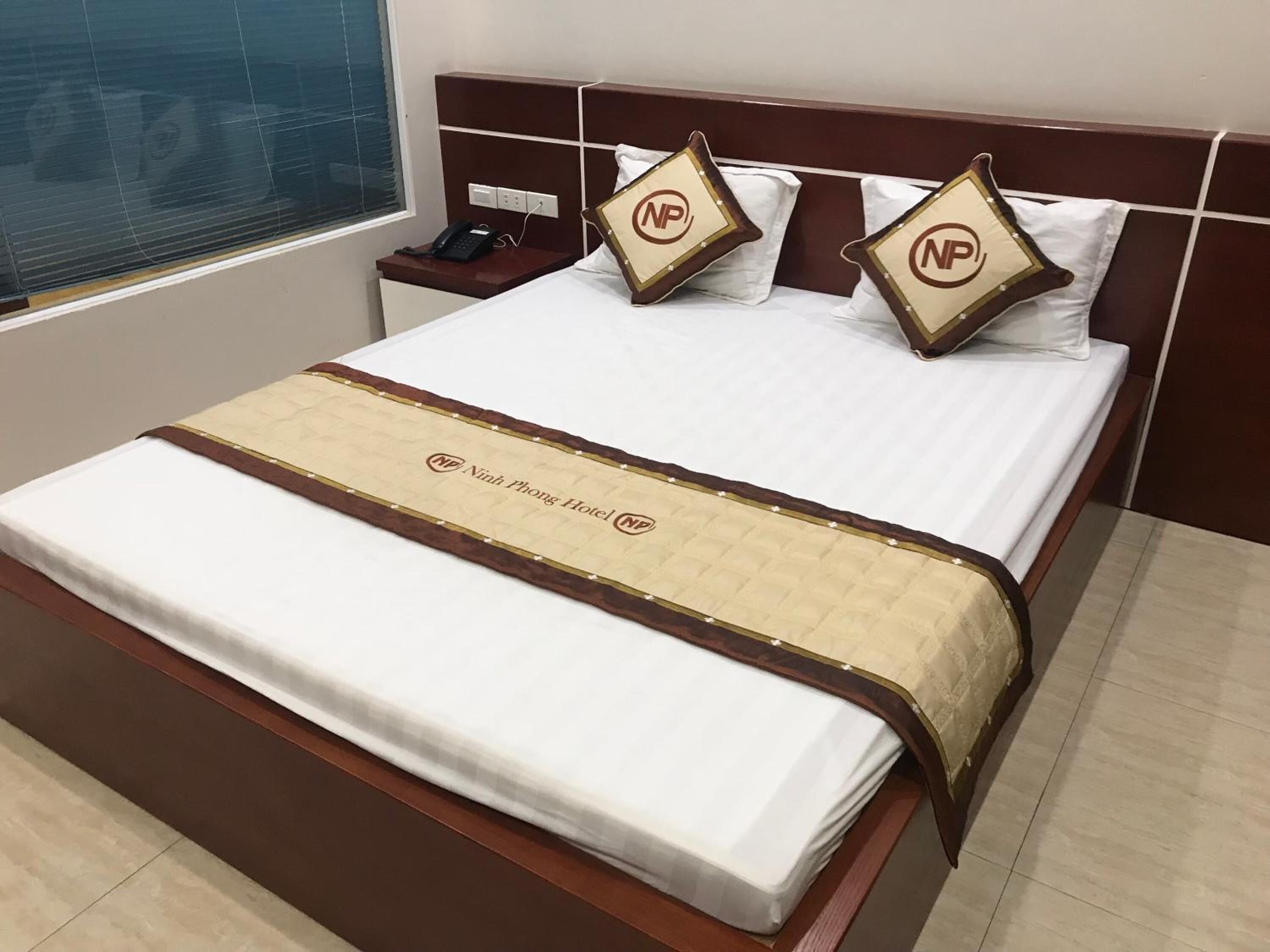A hotel room with a neatly made bed, a bedside phone, and a window with blinds.