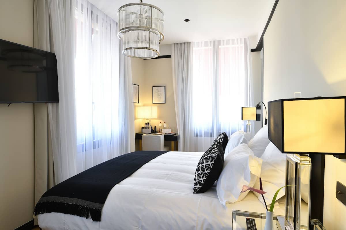 A hotel room with a bed, a TV, a desk with a lamp, and a large window with curtains.