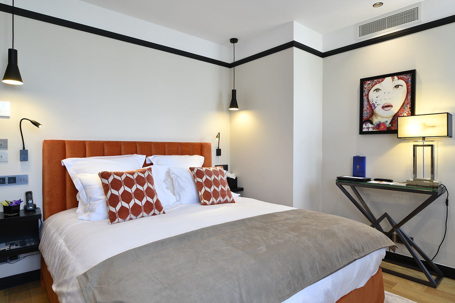 A modern hotel room with a large bed, a desk, and a wall-mounted artwork.