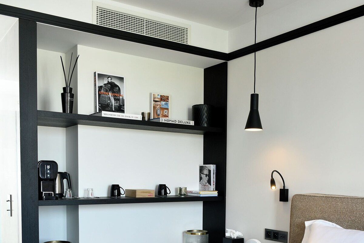 A modern hotel room with a bed, a desk with a computer, shelves with books and decorative items, and a wall-mounted light fixture.