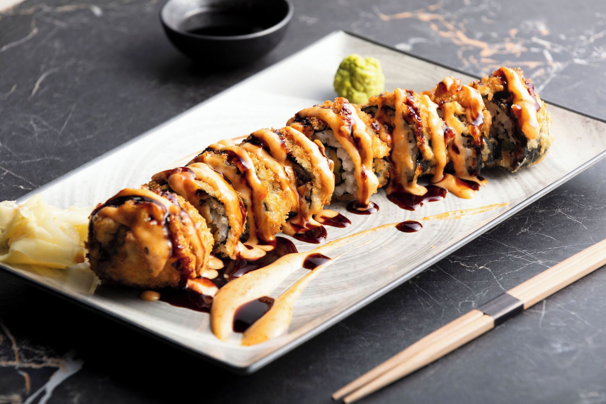 Un plato de sushi sobre una bandeja rectangular, con palillos y un pequeño tazón de salsa.