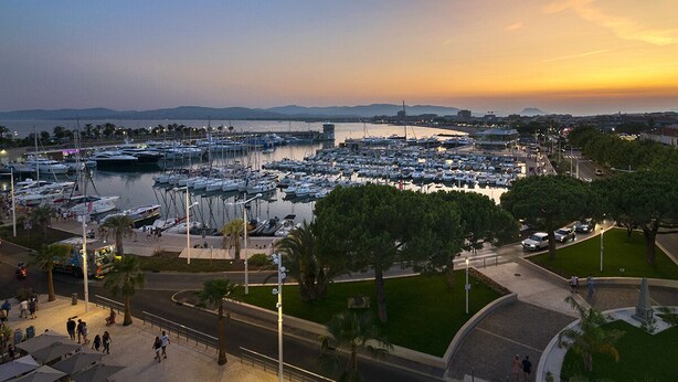 A marina with numerous boats docked, a pedestrian walkway, and a view of the cityscape in the background. - Hôtel Restaurant Le Touring (Saint-Raphael)