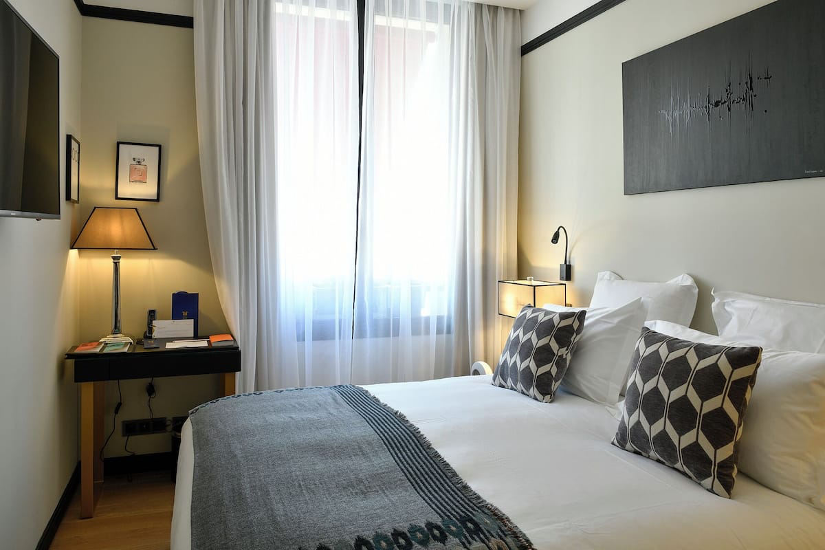 A hotel room with a bed, two bedside tables, a lamp, a painting, and a window with sheer curtains.