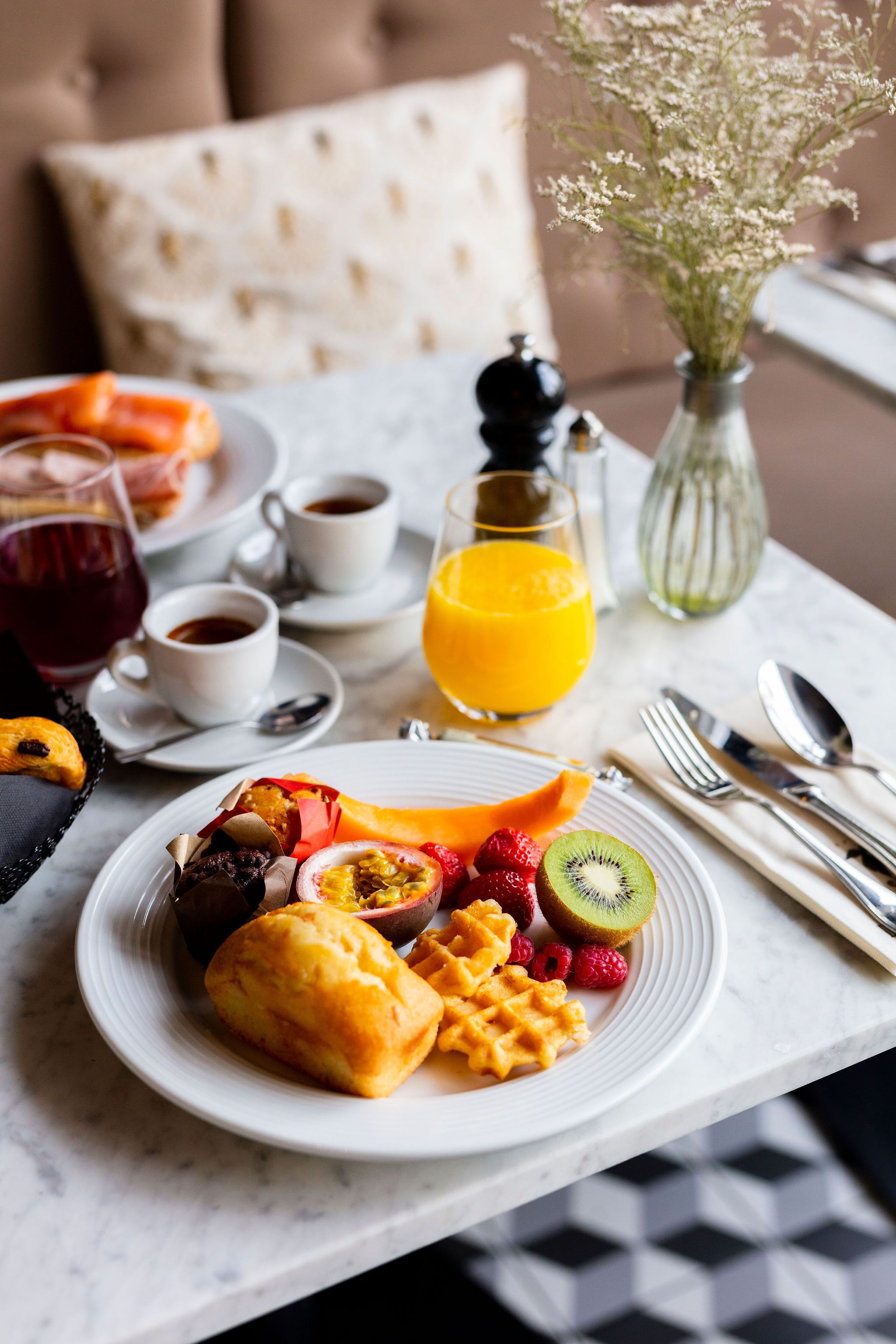 Een ontbijtsetting met een bord wafel, fruit, gebak, een glas sinaasappelsap en een kop koffie op een tafel.